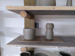 A small cup and small body and head vase in wet clay on a shelf.
