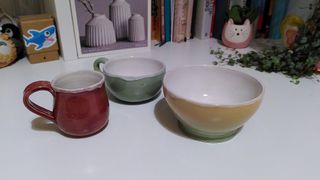 Three finished ceramic vessels: a dark red bulbous cup with a flat handle, a light green cup and a green to yellow gradient bowl, all white inside with the white overflowing out atop their rims.
