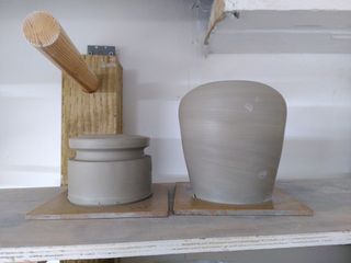 Side shot of two clay forms on their bats after throwing: a table lamp base tapering at the bottom and curving inwards at the top and a closed cylinder with a large groove around the top.