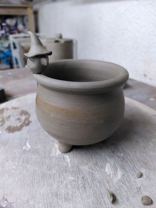 Clay cauldron with a little crow wearing a witch hat on its rim.