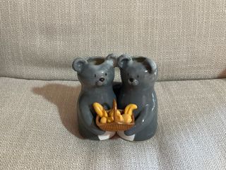 Two vases in the shape of grey bears hugging each other by the shoulder while holding a basket full of bread.