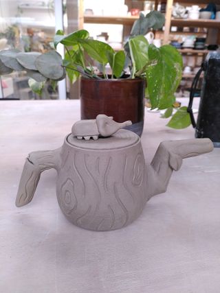 A teapot in dry clay, decorated in stylised tree bark with a branch spout and handle, as well as a lid with rings like the insides of a tree topped by a beetle.