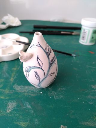 Front picture of a mini bud vase with a mouse poking its nose on the left of its collar, with marker drafting a vine-y pattern on the vase properly drawn in green underglaze. 