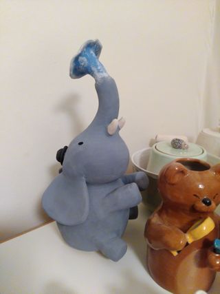 Front ¾ high angle photo of a ceramic sculpture painted in underglaze: a sat-down grey elephant with a black hat spits blue water with white foam from its trunk