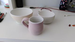 Ceramics coated in glaze sitting on top of a white desk: a light green to yellow gradient bowl, a green cup and a light redish/purple mug, all of them with their inside white and overflowing over the rim.
