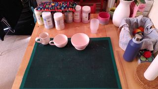 Three bisque fired vessels on a craft mat, with an array of glaze containers behind them, ready for decoration.
