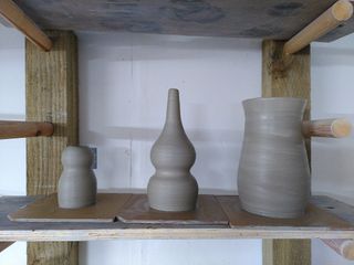 Three clay forms on bats on a shelf waiting to dry: a head and body vase, a double gourd vase and a tall & mildly bulbous vase.
