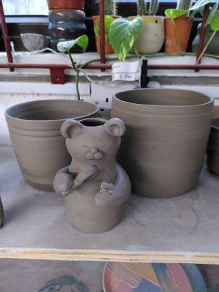 Two planters and a little vase shaped as a bear inspecting a butterfly with a magnifying glass, all leather hard on a shelf.
