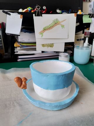 A white pinched pot with its top half painted half blue sits on its saucer, light blue outside and white inside with an orange squirrel figurine sitting on its rim