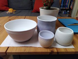 A collection of clay pots painted in glaze: a light-red bowl with white stripe and inside, a textured blue and light-red planter, a yellow cup with blue inside and shade at the rim, and a white jar with a light green lid blending in the body.