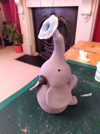 Back ¾ high angle photo of a ceramic sculpture painted in underglaze: a sat-down grey elephant with a black hat spits blue water with white foam from its trunk