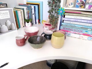 A bunch of ceramics set on a white desk: a red bulbous cup with a flat handle, a red bowl, a green bowl with a dark inside, a blue mug, a pink one, a green and yellow planter, and a little ghost vase.
