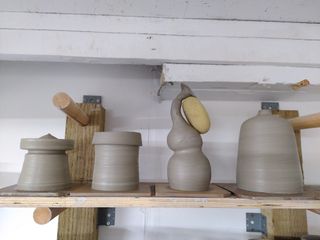 Four clay vessels drying on their bats on a shelf: a circus tent shaped closed form with an indent under the top, a tower like form, and elephant shaped double gourd form and a tapered cylinder closing into a small opening.