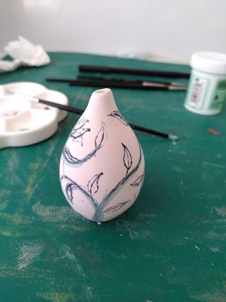 Side picture of the same mini bud vase with marker drafting a vine-y pattern on the vase properly drawn in green underglaze.