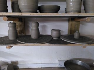 Thrown clay vessels drying on their bats: two small character shaped vases, a cauldron body and a tiny bottle-shape bud vase.
