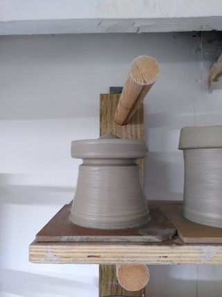 Close up of a clay vessel drying on a shelf: a circus tent shaped closed form with an indent under the top