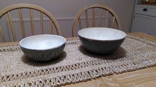 Two serving bowls on a table both with a whiteish inside. Left one's outside is carved with organic strokes and glazed in a glossy blue. Right one's outside is flat with a dark blue-green matt glaze.