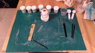 A craft mat with pots of underglaze and brushed laid on it, next to a small vase shaped as a lion king wearing a mantle and crown