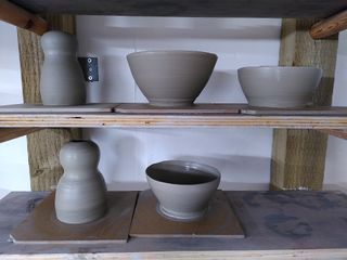 Wet stoneware forms waiting to dry on their bats after throwing. Top shelf holds a 'head and body' vase and two bowls. Bottom shelf a 'head and body' vase.
