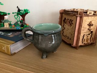 A black cauldron mug supported by three feet and glazed light blue-green inside, overflowing heavily at the rim.