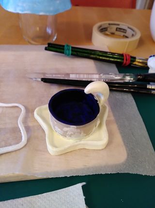 A freshly painted night themed tea light holder. The tea light container painted night blue is still covered by tape, protecting it from the yellowish paint covering a star underneath it and an moon on its side'