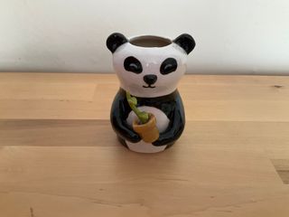 Little vase in the shape of a panda, holding a yellow pot where a green plant with two leaves is growing.