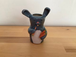 A little vase in the shape of a grey rabbit holding a bunch of carrots and ready to eat one.