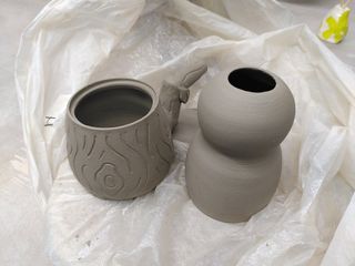 A leather hard teacup with its outside carved in a stylised wooden texture, next to a leather hard vase on top of a plastic sheet.
