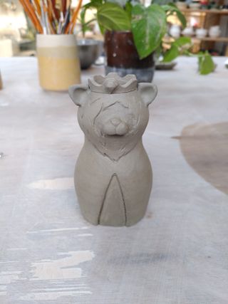 An in progress shot small clay vase shaped as a crowned lion wearing a mantle. The crown holds a large gemstone at the front and the beard and mantle are created as a relief sculpture against the surface of the vase. 
