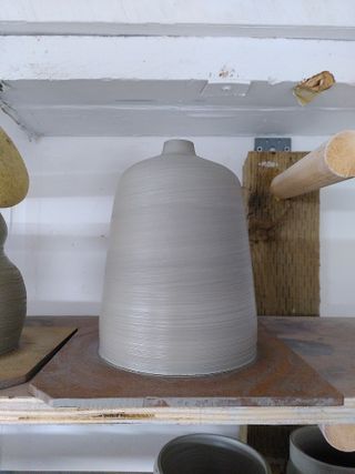 A tapered cylinder clay vessel, closing at the top into a small opening, drying on a shelf.