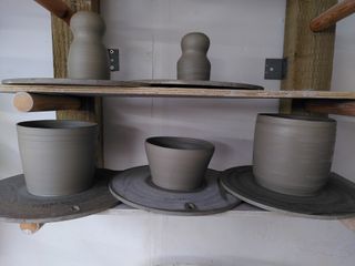 Trown clay forms waiting to dry on their bats. Top shelf has two 'head and body' vase (right one slightly shorter). Bottom shelf has three planters, two bulky ones on the side (with a stripe carved towards the top) and a shorter one in the middle.
