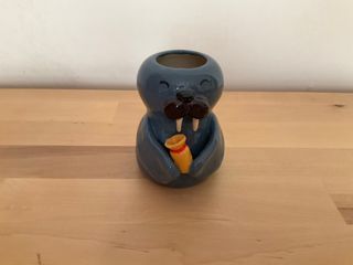 A little vase shaped like a grey-blue walrus holding a yellow vase with a red band.