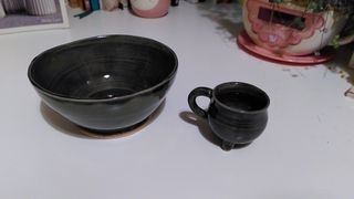 A dark green bowls with black inside next to a small cauldron-shaped cup.
