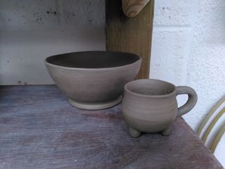 A trimmed bowl and cauldron shape cup in leather hard stoneware clay sitting on the corner of a shelf.
