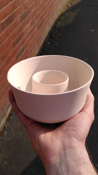 A hand holding a cup stacked inside a bowl, both bisque fired