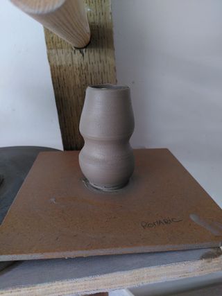 A little wonky vase of wet clay, with a round base surmounted of a slightly conic shape.
