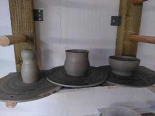 Three clay forms waiting to dry on their bats on a shelf: a 'body and head' vase, a bulbous small planter and a bowl.
