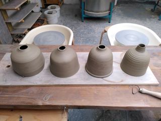 Leather hard bedside lamps bases after trimming, some of them with loose carvings on them. Forms have straight sides curving into a shallow neck where the bulb would go.