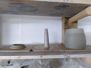 Three thrown clay pieces drying on their bat: a lid, a tall and narrow cone and a curvy pot.
