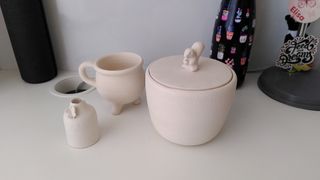 Three bisque fired ceramic vessels on a white table: a mini bottle-shaped bud base with a butterfly on its colar, a cauldron shaped cup and a curvy pot with a squirrel sitting atop its flat lid.
