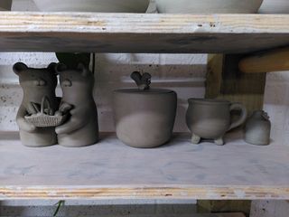 A collection of leather hard ceramic pieces on a shelf: a bear-shaped double vase with two bears holding each other and a basket of bread, a curvy pot with an inset flat lid surmounted by a squirrel handle, a cauldron shaped mug, a bottle shaped mini bud vase with a butterfly.
