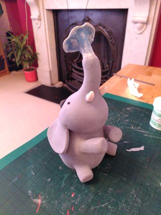 Front ¾ high angle photo of a ceramic sculpture painted in underglaze: a sat-down grey elephant with a black hat spits blue water with white foam from its trunk
