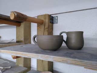 Two leather hard cups waiting to dry further on a shelf: one round with a round handle and one narrow and bulbous with a flat handle.
