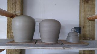 Two clay table lamp bases, angling outwards up before rounding to a flat top with a small hole. Next to them is a little lidded conic jar.