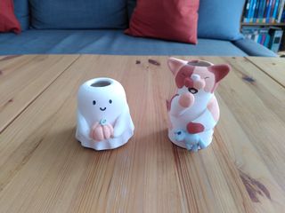 Two small vases coated in underglaze: one in the shape of ghost holding an orange pumpkin with a green stem; the other in the shape of a sleep calico cat with brown and orange spots, holding a light blue teddy-mouse.
