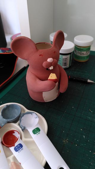 A vase shaped as a mouse painted with a brown body, white belly, red eyes, pink snout and white teeth. It's about to eat a piece of cheese painted yellow with a red rind.
