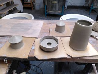 Clay forms waiting to dry on their bats after being thrown: two tiny hut shapped forms, a kind of saucer with a hidden foot and a cone.
