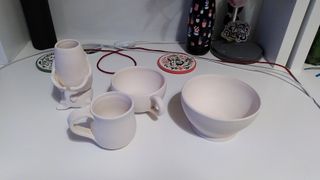 Bisque fired ceramic pieces on top of a desk: a vase shaped as a sleeping bat, two cups and a bowl.
