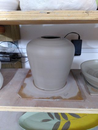 A clay form on a bat after being thrown. The form angle outwards, up from the bottom before rounding inwards to a flat-ish top with a hole.