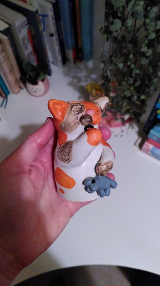 A vase shaped as a sleepy calico cat holding a plush teddy-mouse. Orange spots are very bright and brown spots are washed out, with part of the glaze bubbling.
